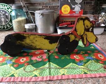 May include: A vintage wooden pull toy dachshund with black and yellow painted details. The toy has a red collar and a black and white face with a smiling mouth. The toy is on a green and pink floral fabric.