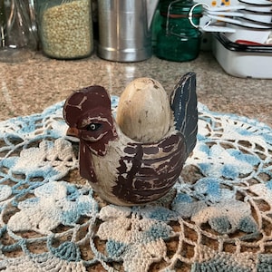 May include: A decorative, hand-painted rooster-shaped egg holder. The rooster is brown, white, and black, with a red comb. It holds a light-coloured egg. The holder sits on a blue and white crocheted doily.