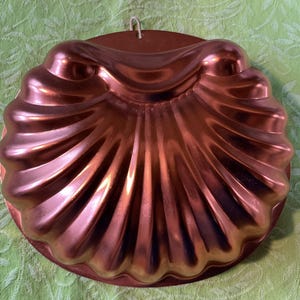 May include: A copper-colored, shell-shaped mold with a scalloped design. The mold has a circular base and a loop for hanging. The surface reflects light, highlighting the curves and ridges of the design. A vintage kitchenware item.