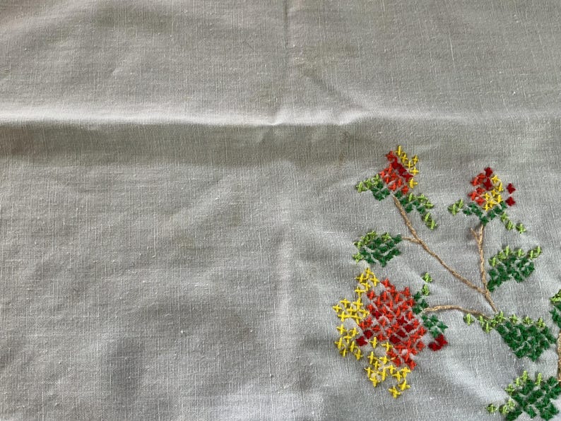 Puede incluir: Tela de lino gris claro con un dise&ntilde;o de bordado floral a punto de cruz. El bordado presenta flores y hojas rojas, amarillas y verdes sobre un tallo marr&oacute;n. La tela tiene una textura sutil.