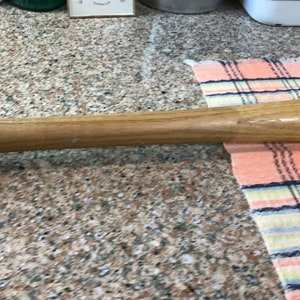 Pu&ograve; includere: Una mazza da baseball in legno con finitura naturale. La mazza &egrave; appoggiata su un piano di lavoro con un tessuto a quadri sullo sfondo.
