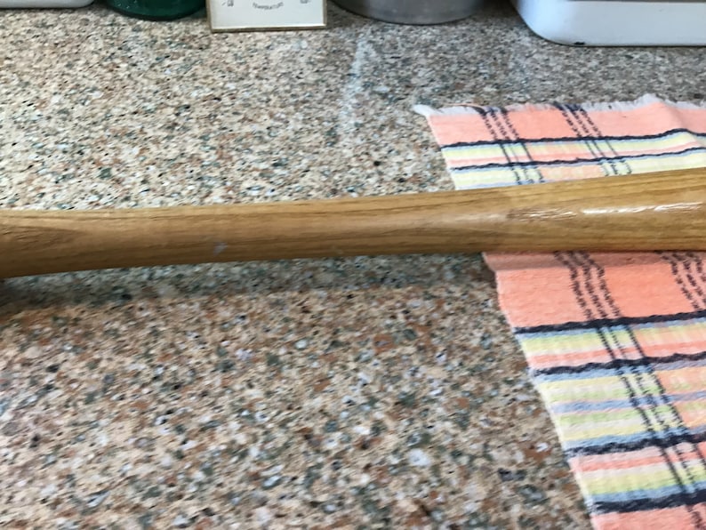 Pu&ograve; includere: Una mazza da baseball in legno con finitura naturale. La mazza &egrave; appoggiata su un piano di lavoro con un tessuto a quadri sullo sfondo.