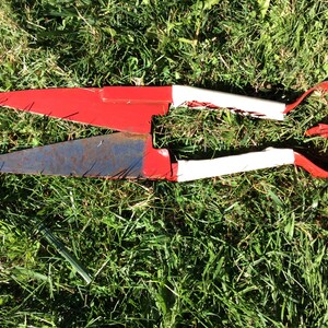 May include: A pair of rusty red and white pruning shears with white handles. The shears are lying on a bed of green grass.