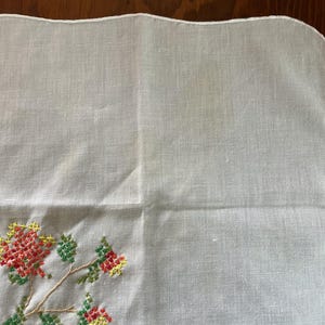 Puede incluir: Pa&ntilde;uelo de lino blanco con borde festoneado. La esquina presenta un dise&ntilde;o floral bordado a punto de cruz en rojo, amarillo y verde. El bordado est&aacute; sobre una rama con hojas y flores.