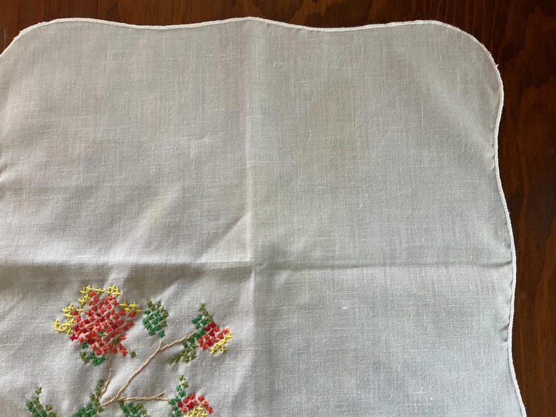 Puede incluir: Pa&ntilde;uelo de lino blanco con borde festoneado. La esquina presenta un dise&ntilde;o floral bordado a punto de cruz en rojo, amarillo y verde. El bordado est&aacute; sobre una rama con hojas y flores.