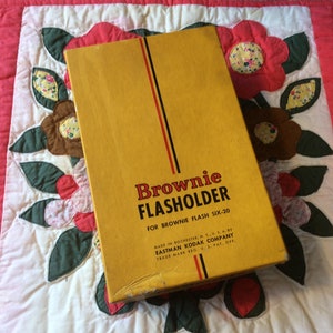 May include: A yellow box with black and red stripes. The box is labeled "Brownie FLASHOLDER for Brownie Flash Six-20" and "Made in Rochester, N.Y. U.S.A. by Eastman Kodak Company Trade Mark Reg. U.S. Pat. Off."