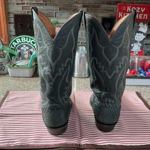 May include: A pair of grey cowboy boots featuring a textured snakeskin-patterned lower section and a smooth leather upper shaft with intricate tonal stitching details. The boots have a tall, pointed silhouette with a stacked heel and tan lining.