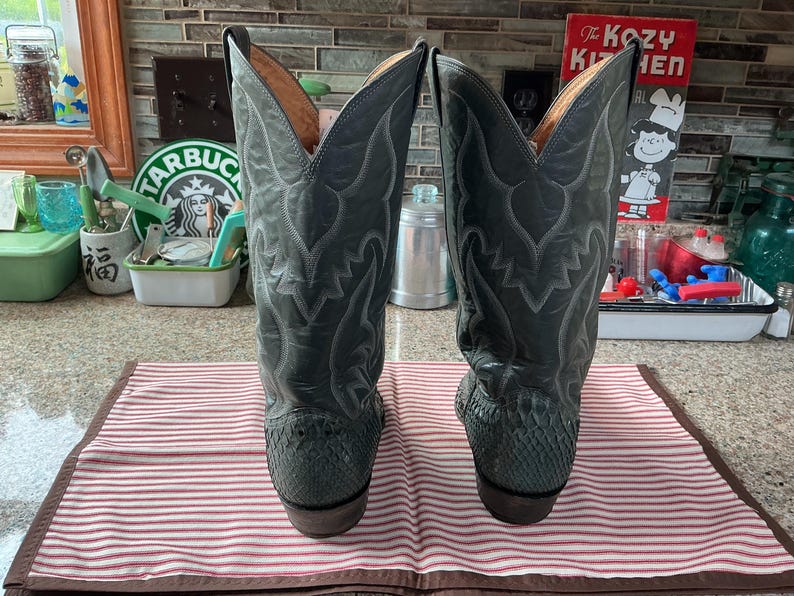 May include: A pair of grey cowboy boots featuring a textured snakeskin-patterned lower section and a smooth leather upper shaft with intricate tonal stitching details. The boots have a tall, pointed silhouette with a stacked heel and tan lining.