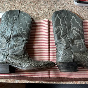 May include: A pair of grey cowboy boots featuring a textured snakeskin-patterned lower section and a smooth leather upper shaft with decorative stitching. The boots have a pointed toe, a stacked heel, and pull tabs at the top of the shaft.