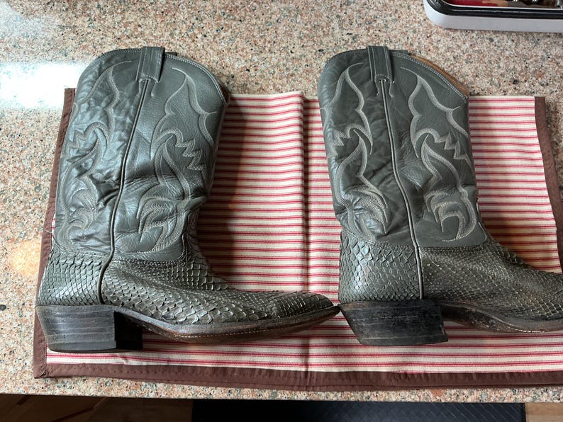 May include: A pair of grey cowboy boots featuring a textured snakeskin-patterned lower section and a smooth leather upper shaft with decorative stitching. The boots have a pointed toe, a stacked heel, and pull tabs at the top of the shaft.