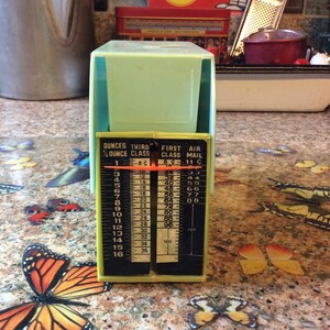 May include: A green and yellow postal scale with a sliding weight indicator. The scale has markings for ounces and grams. The scale is labeled "DUNCES THIRD 1/4 OUNCE CLASS 1-8C" and "FIRST AIR CLASS MAIL 8C-11C".