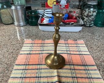 May include: A vintage brass candlestick holder with a round base and a flared top. The candlestick is placed on a plaid orange, yellow, and blue patterned cloth. Other kitchen items are in the background.