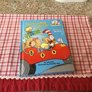 May include: A children's book titled "There's a Map on My Lap!" with an illustration of a red car with a group of children and a cat in a hat driving. The book is part of a series called "All About MAPS".