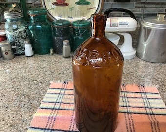 May include: A vintage amber glass bottle with the word "SPRAY" embossed on the front. The bottle is sitting on a checkered cloth.