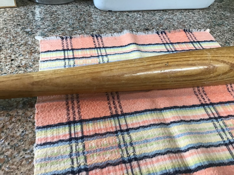 Pu&ograve; includere: Una mazza da baseball in legno con finitura naturale. La mazza &egrave; appoggiata su un panno a quadri rosa e bianco.
