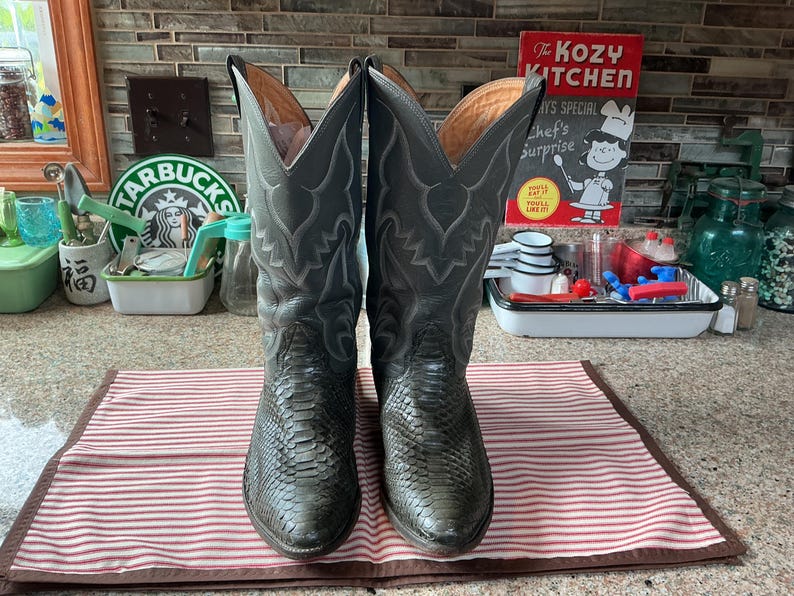 May include: A pair of dark grey cowboy boots featuring a textured snakeskin-patterned leather foot and a tall shaft with decorative tonal stitching. The boots have a pointed toe design and a tan interior lining, standing upright on a striped surface.