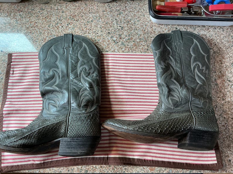 May include: A pair of grey cowboy boots featuring a textured snakeskin-patterned leather foot and a smooth leather shaft with decorative tonal stitching. The boots have a pointed toe, a stacked block heel, and a pull-on design with side tabs.