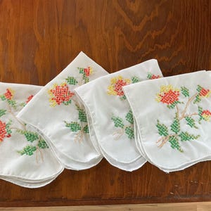 Puede incluir: Cuatro pa&ntilde;uelos blancos bordados con dise&ntilde;os florales. Cada pa&ntilde;uelo presenta un patr&oacute;n floral de punto de cruz en rojo, amarillo y verde. Los pa&ntilde;uelos est&aacute;n dispuestos sobre una superficie de madera.