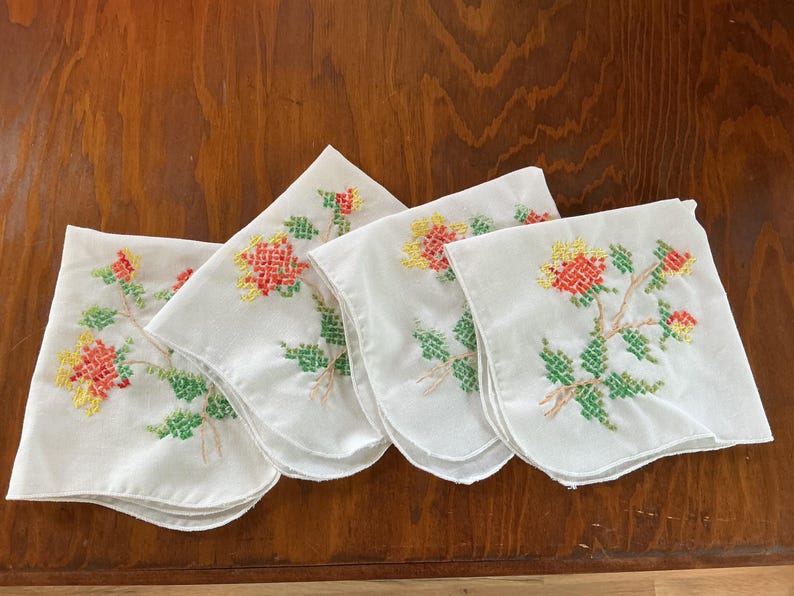 Puede incluir: Cuatro pa&ntilde;uelos blancos bordados con dise&ntilde;os florales. Cada pa&ntilde;uelo presenta un patr&oacute;n floral de punto de cruz en rojo, amarillo y verde. Los pa&ntilde;uelos est&aacute;n dispuestos sobre una superficie de madera.