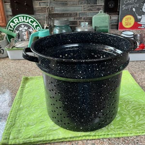 May include: A deep, cylindrical black colander with a white speckled enamel finish. The kitchen tool features two small side handles and a series of drainage holes distributed across the sides and base, designed for straining food items.