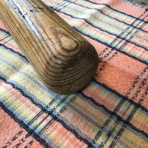 Pu&ograve; includere: Primo piano di una mazza da baseball in legno con una finitura naturale usurata. La mazza &egrave; appoggiata su un tessuto a quadri rosa e blu.