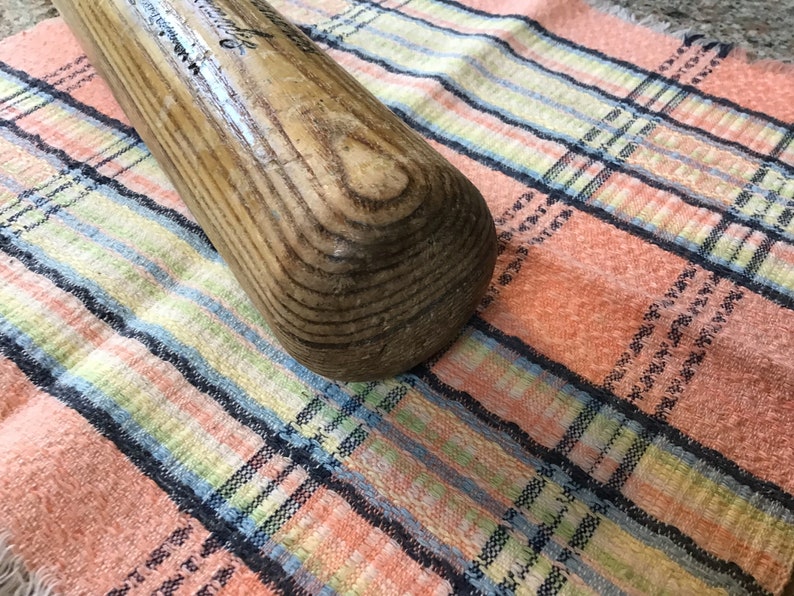 Pu&ograve; includere: Primo piano di una mazza da baseball in legno con una finitura naturale usurata. La mazza &egrave; appoggiata su un tessuto a quadri rosa e blu.