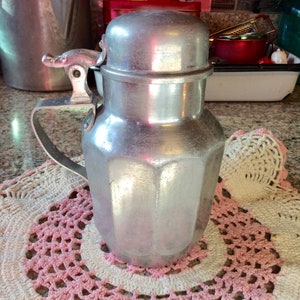May include: A silver aluminum pitcher with a hinged lid and a handle. The pitcher has a textured, octagonal design and is sitting on a pink and white crocheted doily.