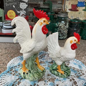 May include: Two white ceramic rooster and hen figurines with bright red combs and wattles, standing on textured green bases. The rooster is larger than the hen, and both feature yellow beaks and feet with detailed feather textures.