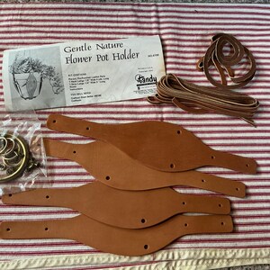 May include: A kit for a flower pot holder, featuring pre-punched leather parts, leather strips, and metal hardware. The kit includes a paper with the text "Gentle Nature Flower Pot Holder". The leather parts are tan.