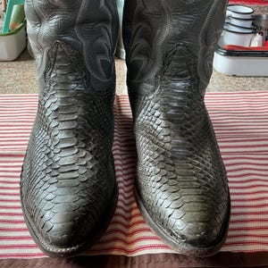May include: A pair of dark grey cowboy boots featuring a textured snakeskin pattern on the vamp and toe, with smooth leather shafts that display decorative stitching. The boots have a pointed toe design and a tall, structured silhouette.