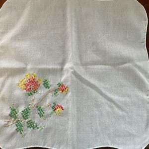 Puede incluir: Un pa&ntilde;uelo blanco cuadrado con un dise&ntilde;o floral de punto de cruz. El bordado presenta flores y hojas rojas, amarillas y verdes sobre un tallo marr&oacute;n. Los bordes del pa&ntilde;uelo tienen un ribete festoneado.