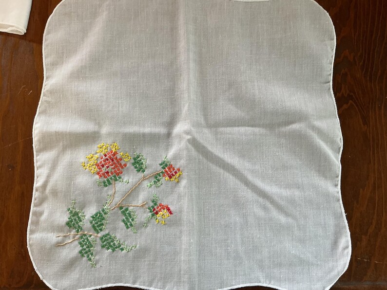 Puede incluir: Un pa&ntilde;uelo blanco cuadrado con un dise&ntilde;o floral de punto de cruz. El bordado presenta flores y hojas rojas, amarillas y verdes sobre un tallo marr&oacute;n. Los bordes del pa&ntilde;uelo tienen un ribete festoneado.