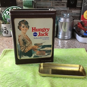 May include: A vintage brown metal tin with a red and white label for Hungry Jack Buttermilk Complete Pancake Mix. The label features a woman in a white dress holding a pan of pancakes. The text "Hungry Jack" is in red and white.