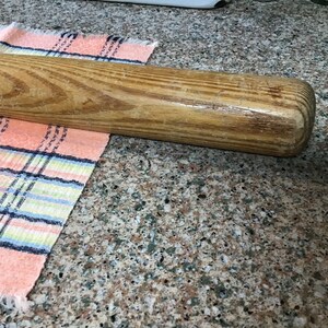 Pu&ograve; includere: Una mazza da baseball in legno con finitura naturale. La mazza &egrave; appoggiata su un piano di lavoro con un panno a quadri rosa e blu.