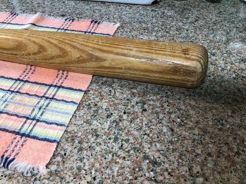 Pu&ograve; includere: Una mazza da baseball in legno con finitura naturale. La mazza &egrave; appoggiata su un piano di lavoro con un panno a quadri rosa e blu.