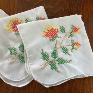 Puede incluir: Un conjunto de pa&ntilde;uelos blancos con dise&ntilde;os florales bordados. Las flores son rojas y amarillas, con hojas y tallos verdes. El bordado es detallado y a&ntilde;ade un toque vintage a los pa&ntilde;uelos.