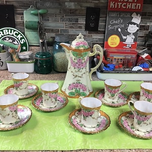 Puede incluir: Un juego de té vintage con una tetera de color crema y seis tazas y platillos a juego. El juego presenta un diseño floral con rosas rosas y hojas verdes, acentuado con ribetes dorados.