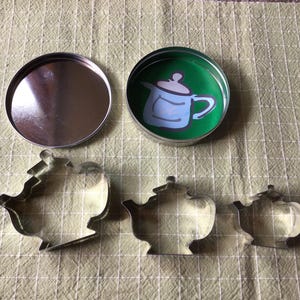 Peut inclure: Un ensemble de trois emporte-pièces en métal en forme de théière, une boîte ronde avec un couvercle et une boîte avec un intérieur vert représentant une illustration de théière bleue. Les articles sont disposés sur une surface à carreaux vert clair.