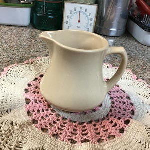 May include: A cream-colored ceramic pitcher with a handle. The pitcher is sitting on a crocheted doily with pink and white accents.