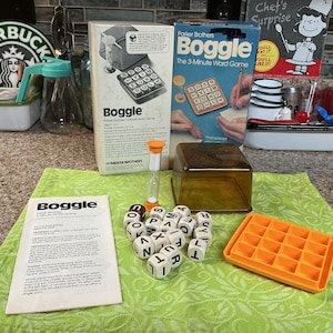 Pode incluir: Um conjunto de jogos de palavras Boggle vintage. O conjunto inclui a caixa do jogo, dados, um cronômetro e uma bandeja de plástico laranja. A caixa do jogo é branca e azul com o nome do jogo e instruções.