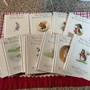 Puede incluir: Una colección de libros de Beatrix Potter, incluyendo "El cuento de Peter Rabbit" y "El cuento de Benjamin Bunny". Los libros tienen cubiertas blancas con ilustraciones de animales y títulos en texto negro. Están dispuestos sobre una tela a cuadros roja y blanca.