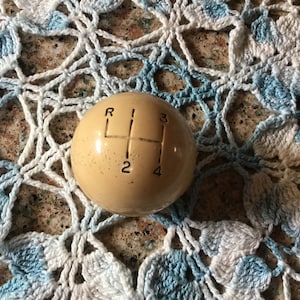 May include: A beige round gear shift knob with black numbers and letters. The knob has the numbers 2, 3, and 4 and the letter R.