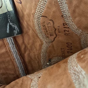 May include: The interior of a tan leather boot featuring a stamped Nocona Boots logo, the word Style, and the numbers 7400 and 8217. The leather has a textured surface with light-coloured decorative stitching patterns visible along the side panels.