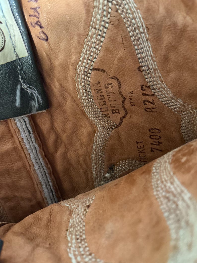 May include: The interior of a tan leather boot featuring a stamped Nocona Boots logo, the word Style, and the numbers 7400 and 8217. The leather has a textured surface with light-coloured decorative stitching patterns visible along the side panels.
