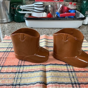 May include: Two glossy brown cowboy boot-shaped planters are displayed on a plaid towel. The planters have a classic western design with a pointed toe and a curved top. Kitchen items are in the background.