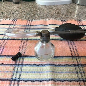 Puede incluir: Un atomizador de vidrio vintage con una botella de vidrio transparente y un bulbo de goma negro. El atomizador tiene un tubo de plástico transparente conectado al bulbo.