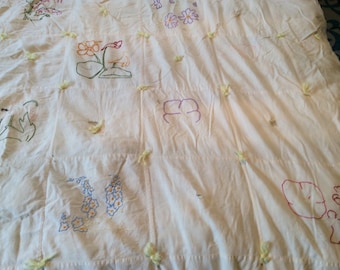 Op de afbeelding: Een wit katoenen quilt met handgestikte borduursels van verschillende bloemen en bladeren. De quilt is vastgemaakt met geel garen.