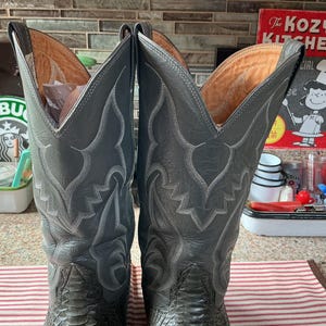 May include: A pair of tall grey leather cowboy boots featuring intricate white stitching patterns on the shafts and a textured, scale-like finish on the lower foot sections. The boots have a high, pointed collar design and tan interior lining.