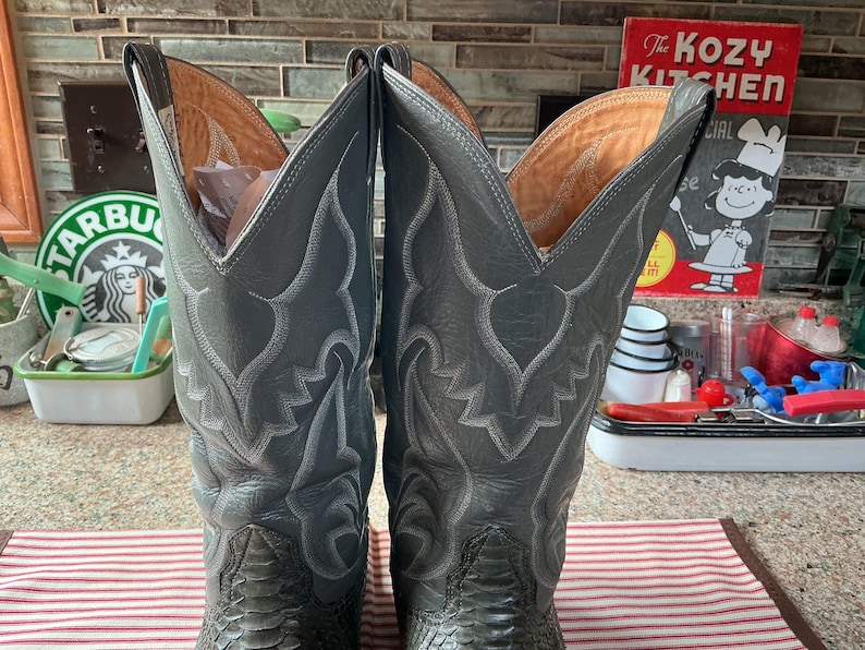 May include: A pair of tall grey leather cowboy boots featuring intricate white stitching patterns on the shafts and a textured, scale-like finish on the lower foot sections. The boots have a high, pointed collar design and tan interior lining.