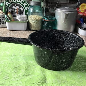 Pode incluir: Uma panela de esmalte preto com acabamento branco salpicado e uma alça preta. A panela está sobre uma toalha de cozinha verde com padrão.